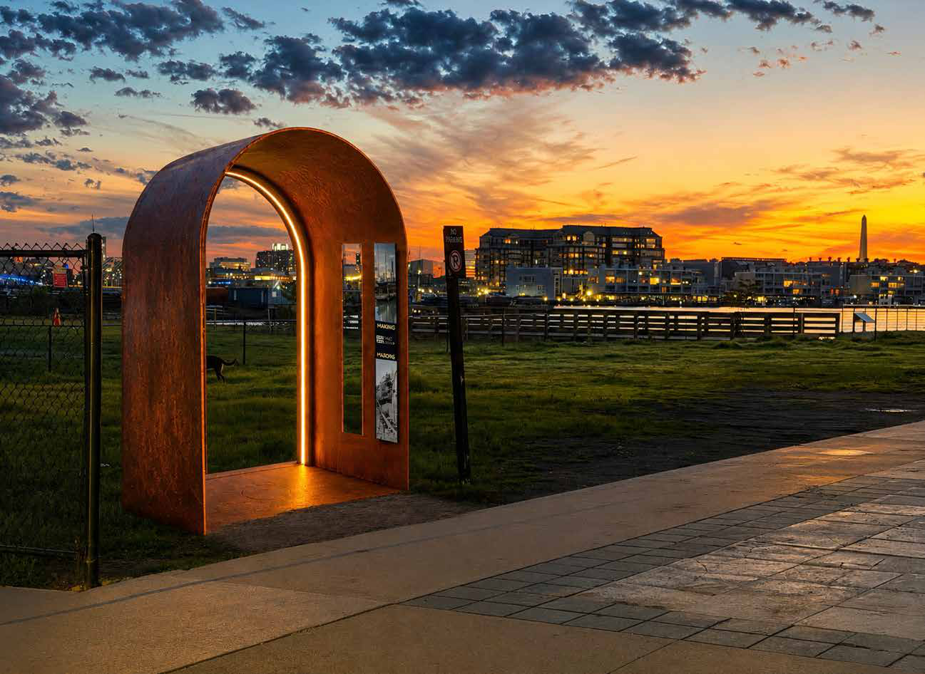 The Thresholds arch, a weathered-steel archway glowing at sunset beside the greenway path, with Boston Harbor and the skyline behind it