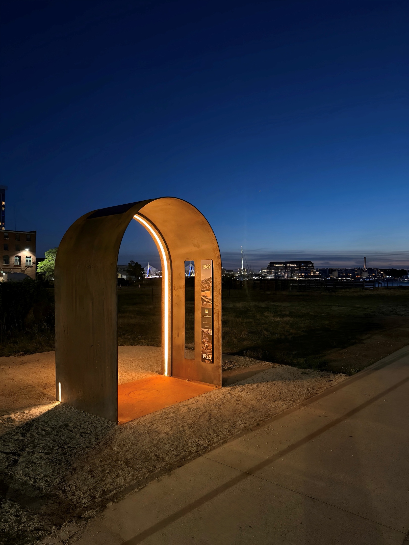 The Thresholds arch, a weathered-steel archway glowing at night at the Atlantic Works Shipyard site, its lit story panel beneath a deep-blue sky with the harbor skyline beyond